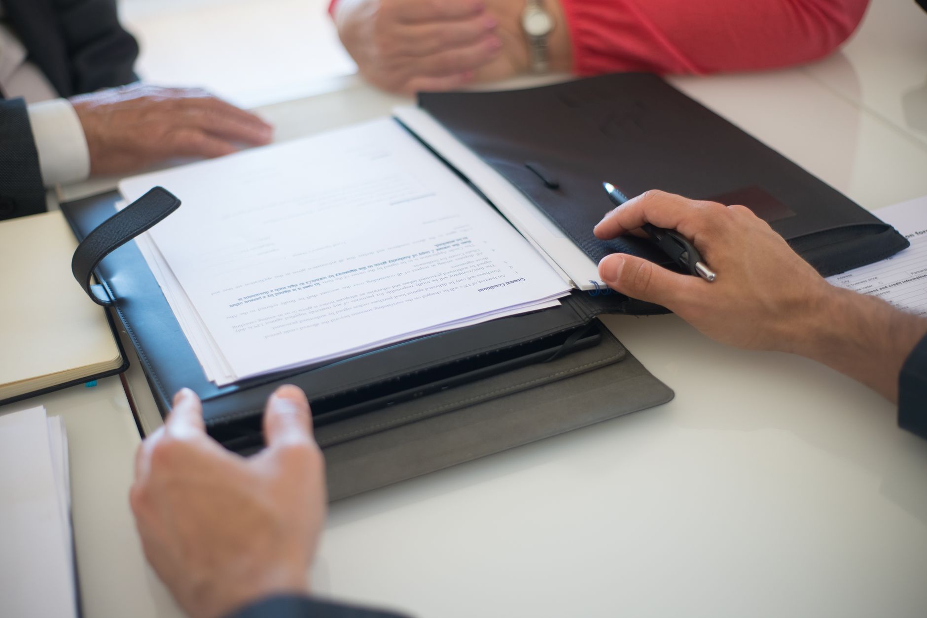 Mains de plusieurs personnes autour d’une table avec une chemise noire contenant des documents et un stylo, lors d’une réunion.